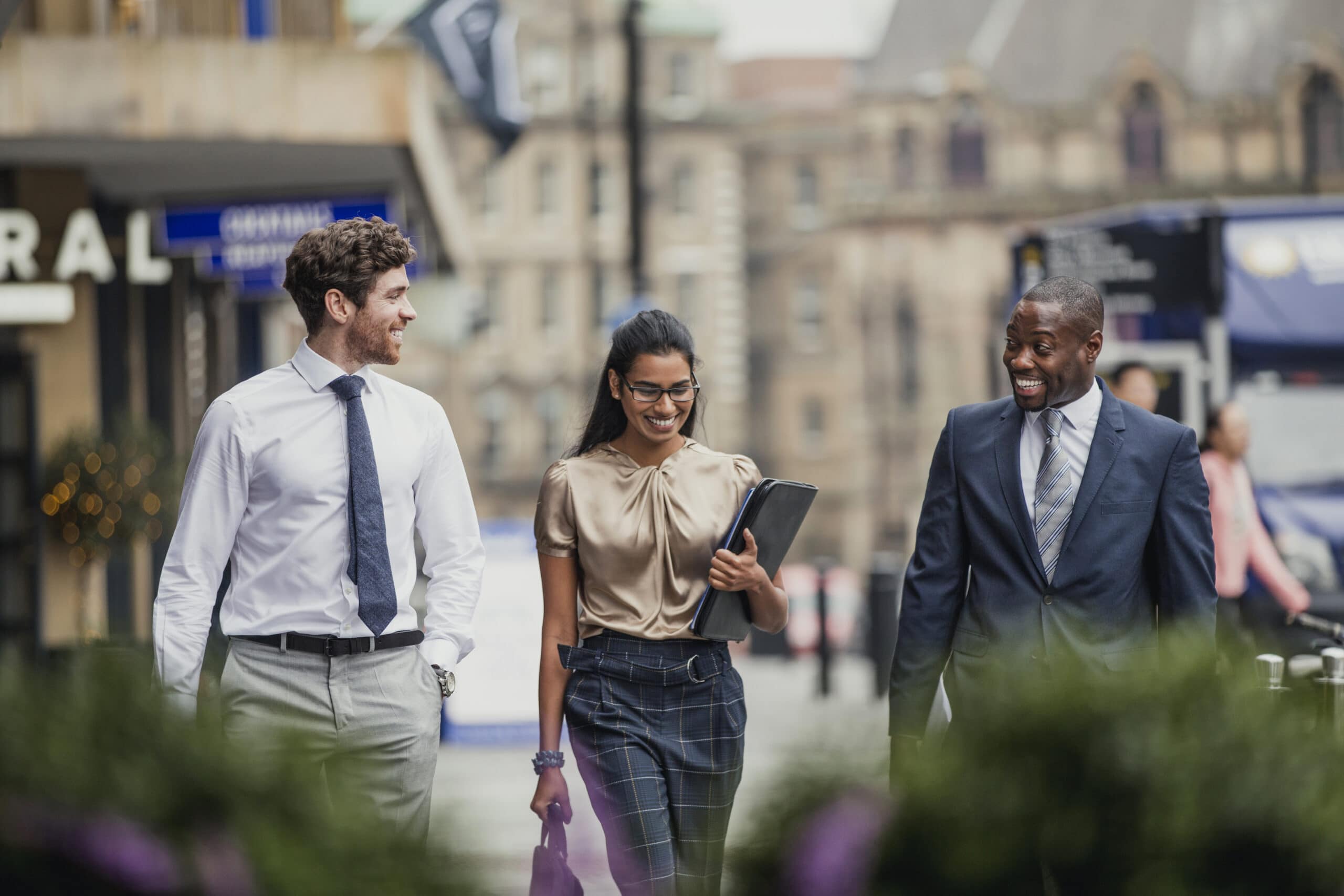 Front view of three business people walking through the city streets. All three people are wearing formal clothing.