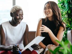 Two professional women communicating over a piece of paper. they are smiling and working collaborating in a casual office environment