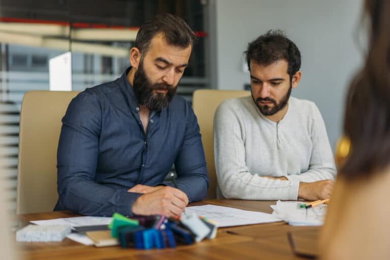 Two male colleagues sit side by side in meeting, reviewing paperwork