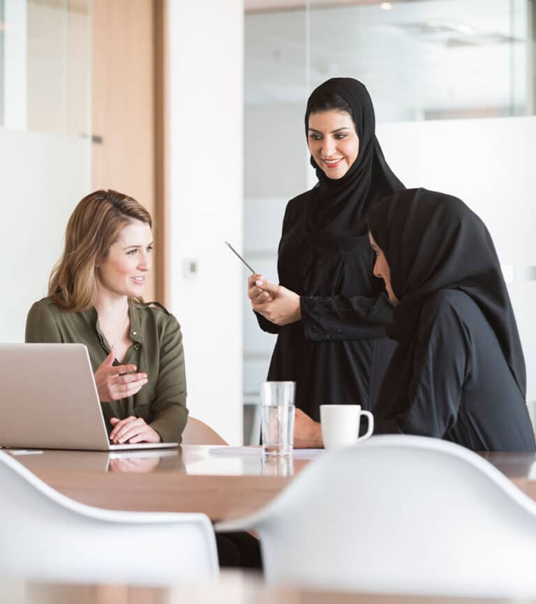 A female sits with her Middle Eastern female colleagues at a desk with a laptop open in conversation