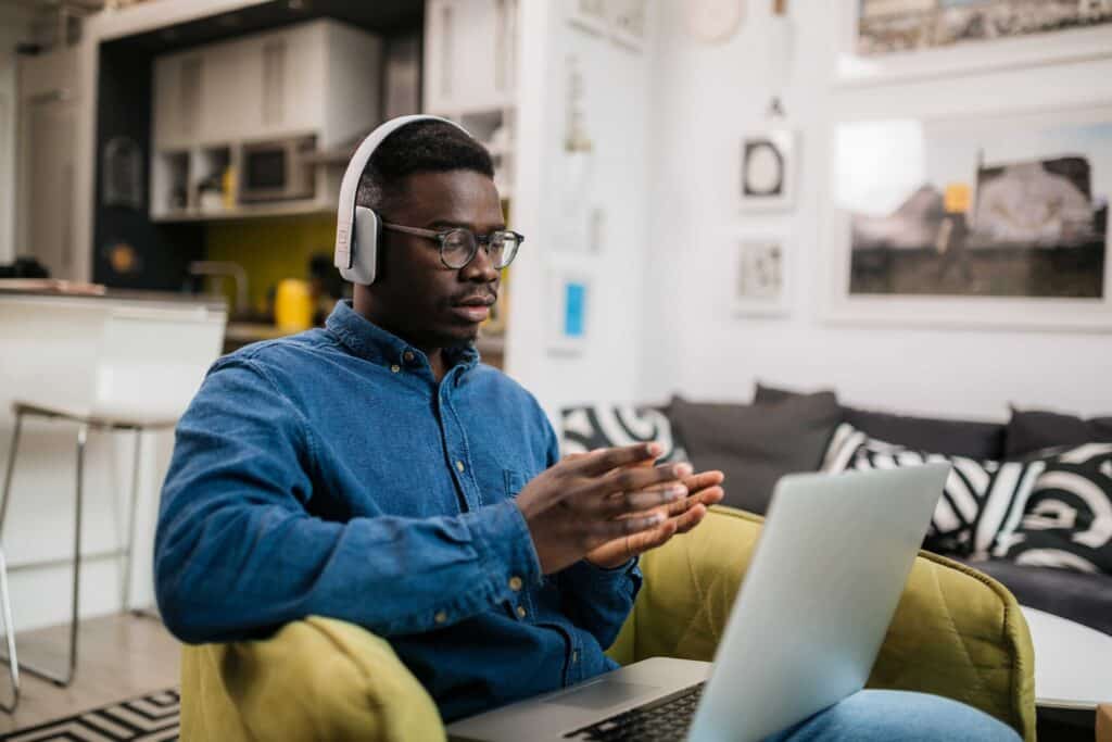 Male has his headphones on and using a laptop to study remotely from home, gesturing with his hands while he's having a conversation with someone using a video call