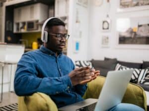 Male has his headphones on and using a laptop to study remotely from home, gesturing with his hands while he's having a conversation with someone using a video call