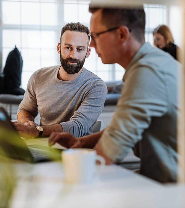 two male colleagues sit at a desk and laptop discussing work