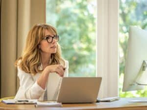 Cheerful smiling mid aged businesswoman sitting at desk and using laptop and computer for work. Confident professional female wearing white blazer and glasses.