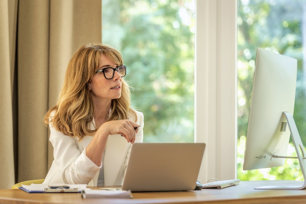 Cheerful smiling mid aged businesswoman sitting at desk and using laptop and computer for work. Confident professional female wearing white blazer and glasses.