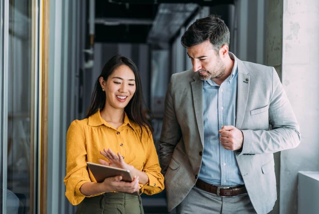 Shot of a two confident business persons talking in the work place. Two colleagues using a digital tablet while walking in a modern office. Businessman and businesswoman in meeting discussing business strategy. Business coworkers working together in the office.