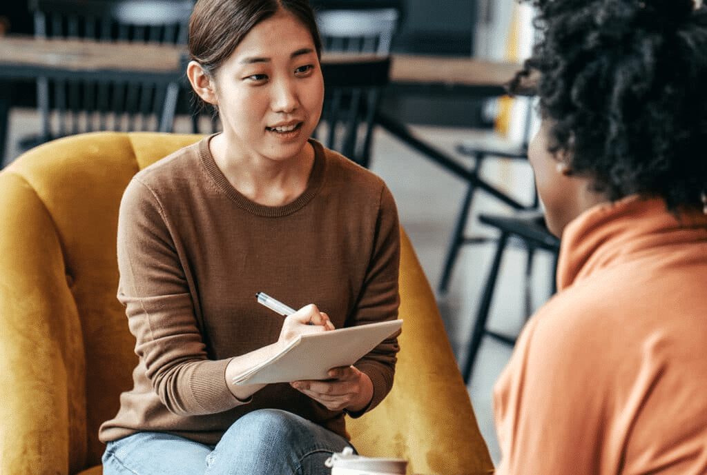 Two women sit chatting, one interviewing the other, with a notepad and pen