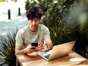 Man working from coffee shop. He is using mobile phone and laptop